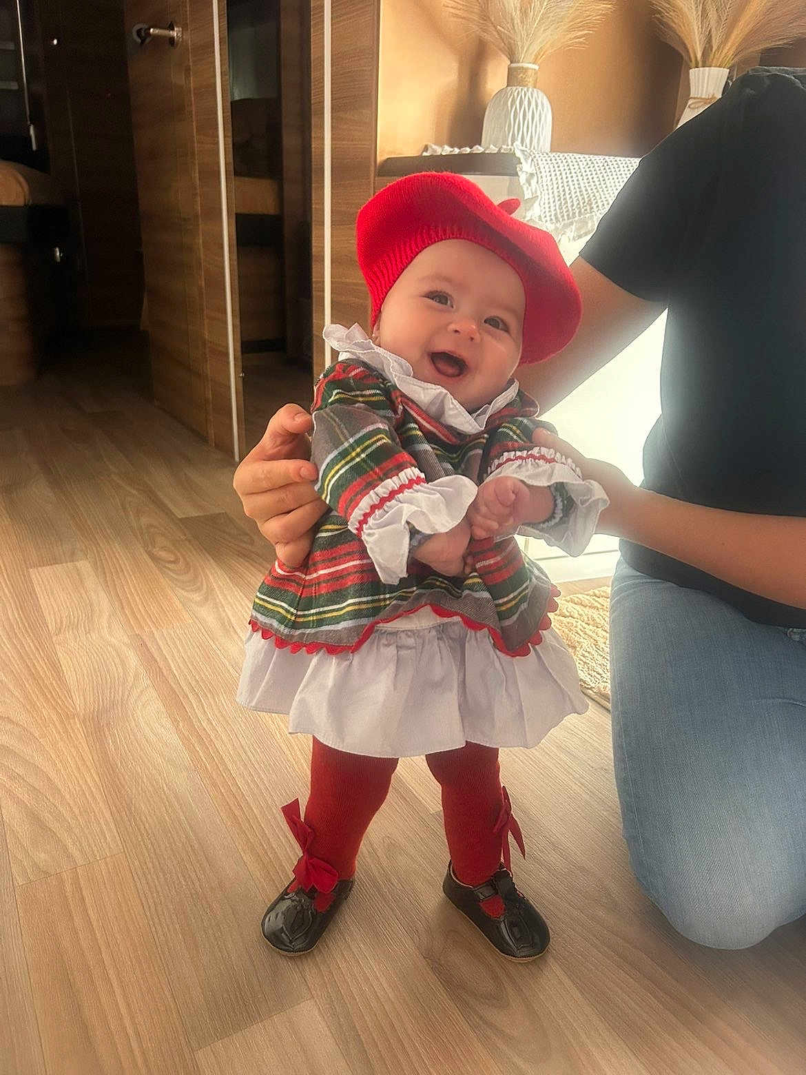 Lïya participe au concours pour gagner de l'argent avec cette photo : baby, child, smiling, red_beret, plaid_dress, indoor, wooden_floor, adult_hand, supporting, happy, footwear, red_tights, person, flooring, home_decor, vase, plant, kneeling, cute, portrait