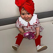 Lïya participe au concours pour gagner de l'argent avec cette photo : toddler, child, red_turban, red_stockings, white_collar, floral_dress, toy, pink_toy, couch, smiling, sitting, indoors, cute, happy, face, legs, footwear, white_shoes, playful, portrait
