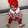 toddler, child, red_turban, red_stockings, white_collar, floral_dress, toy, pink_toy, couch, smiling, sitting, indoors, cute, happy, face, legs, footwear, white_shoes, playful, portrait
