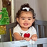Penelope is registered to the contest to win money with this photo: baby, child, toddler, smiling, portrait, curly_hair, hair_bow, face, eyes, teeth, shirt, heart, hands, crib, furniture, indoor, home, laundry_bottle, wood, photography