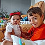 baby, blanket, boy, child, couch, family, fruit_bowl, furniture, happy, home, indoor, orange_shirt, picture_frame, plant, portrait, red_bow, seated, smiling, table, toy