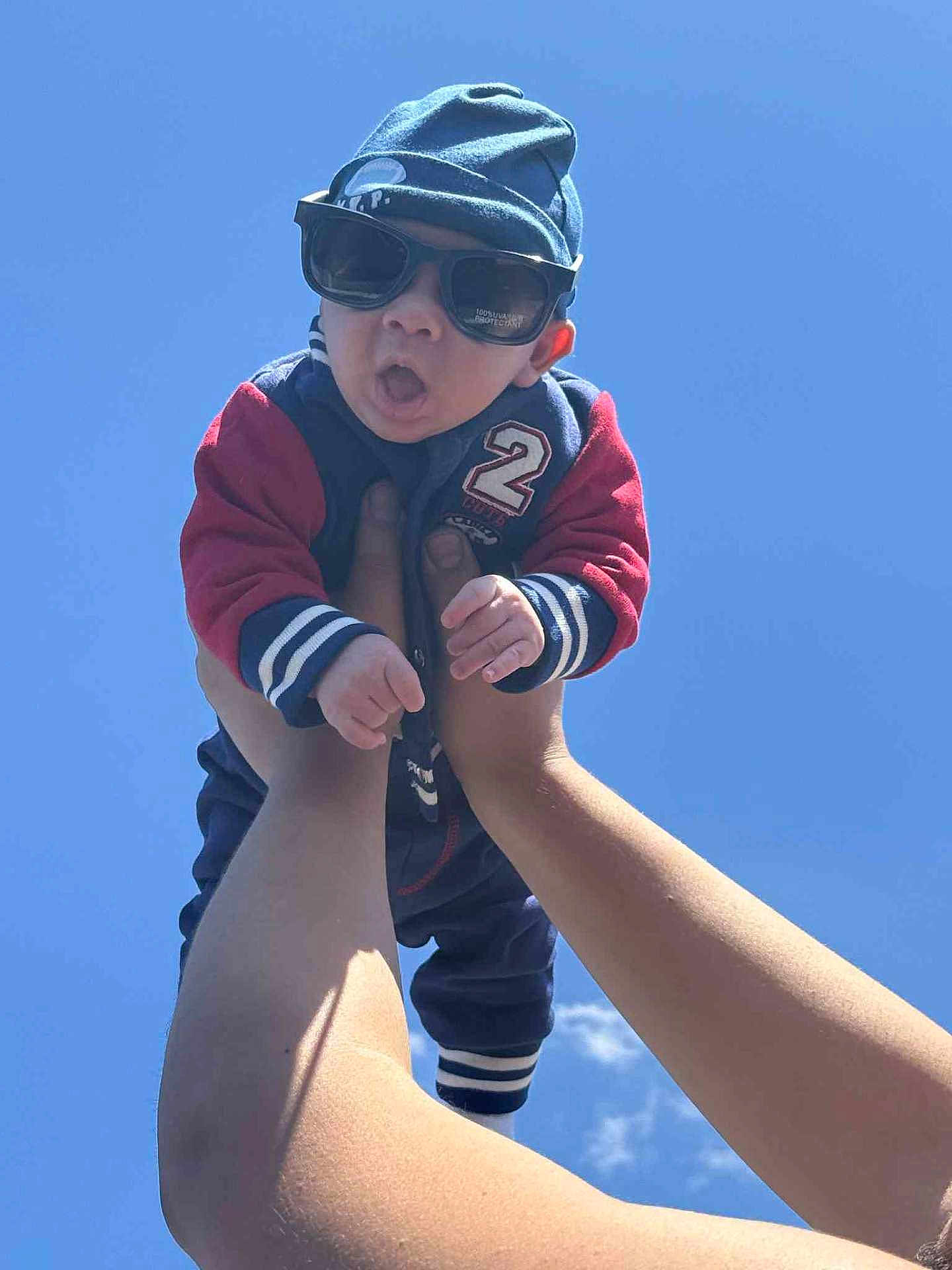 Elias is registered to the contest to win money with this photo: baby, child, sunglasses, hat, blue_sky, arms, hands, jacket, outdoor, person, cute, expression, sunny, casual_clothing, funny_face, infant, headwear, daylight, fashion, portrait