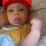 Elias is registered to the contest to win money with this photo: baby, infant, face, tongue, red_hat, mario_cap, blanket, arm, person, clothing, cute, expression, indoors, human, headwear, child, young, wrapped, holding, soft