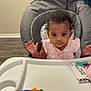 baby, high_chair, pink_clothing, unicorn_print, toy, plush_toy, indoor, wooden_floor, baby_girl, curly_hair, face, hands, tray, expression, child, person, room, flooring, furniture, household