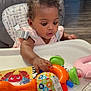 baby, child, high_chair, toy, colorful, curly_hair, indoor, wood_floor, infant, play, person, furniture, pink, orange, green, table, expression, focus, seat, home