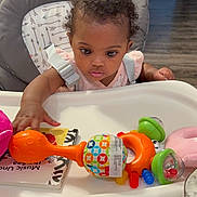 Rose-Shaina joined the competition — help win amazing prizes! baby, high_chair, toy, infant, indoor, curious, child, colorful, plastic_toy, face, person, seat, clothing, table, floor, wood_floor, hand, expression, grip, background