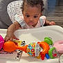baby, high_chair, toy, infant, indoor, curious, child, colorful, plastic_toy, face, person, seat, clothing, table, floor, wood_floor, hand, expression, grip, background