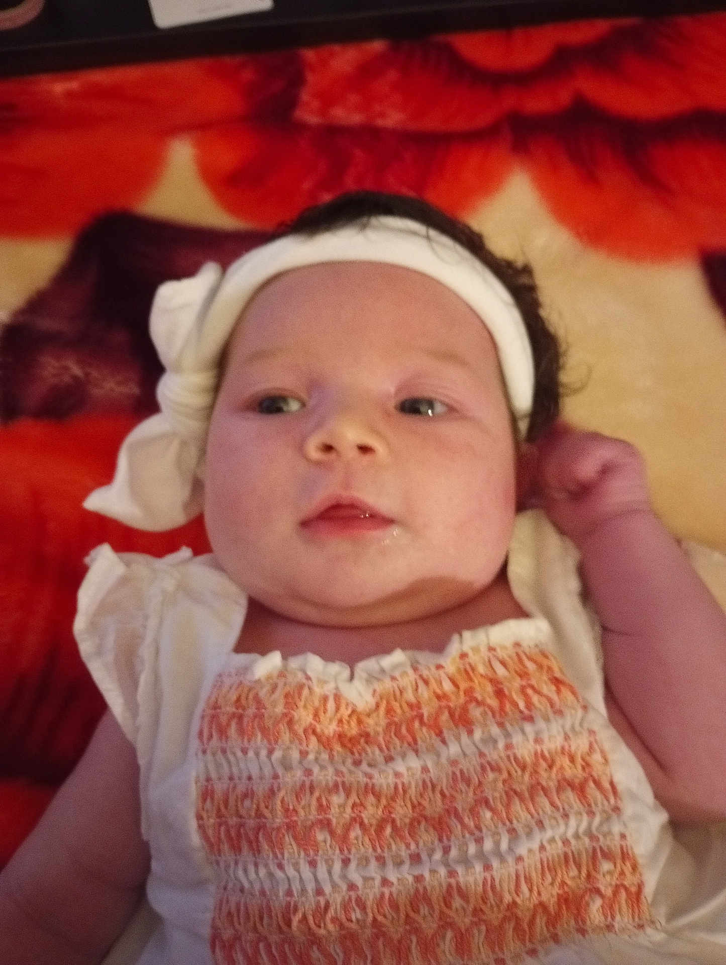 Hazel is registered to the contest to win money with this photo: baby, infant, headband, white_clothing, orange_clothing, blanket, red, orange, face, portrait, child, cute, lying_down, newborn, skin, hand, expression, indoors, soft_texture, young