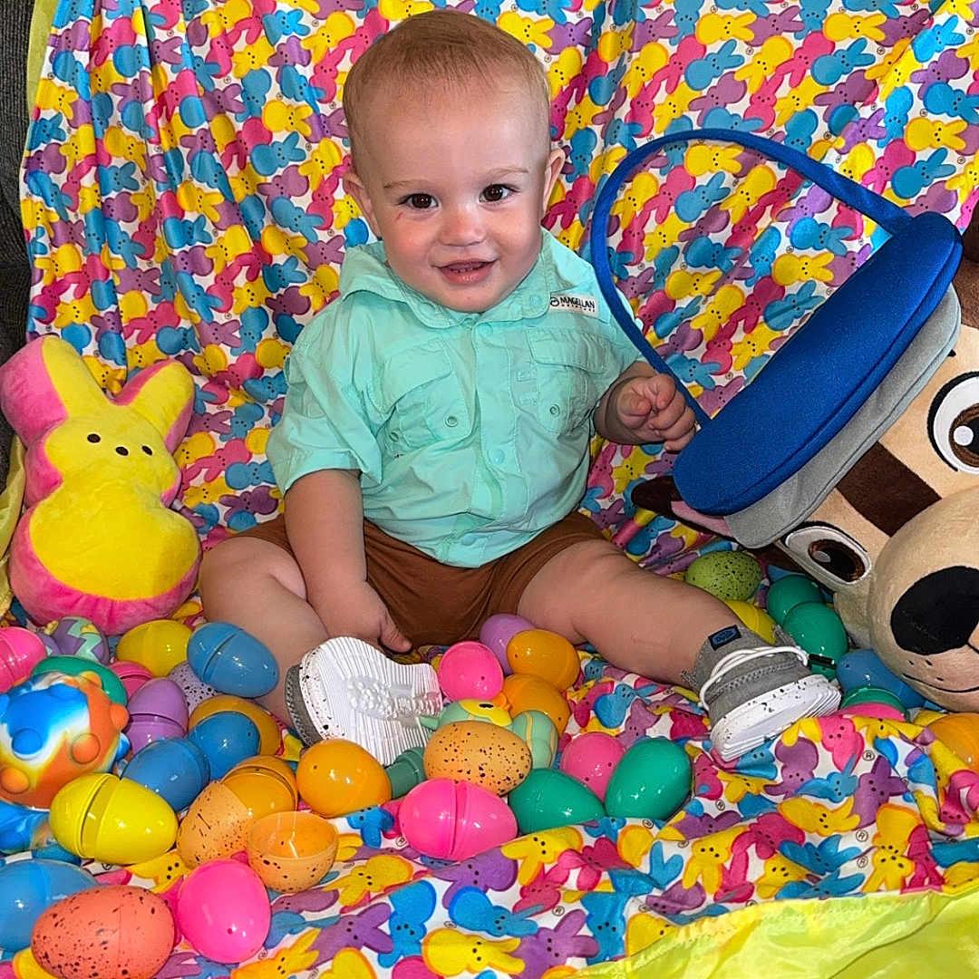 Dallas Wayne joined the competition — help win amazing prizes! blanket, bright, bunny_pattern, child, clothing, colorful, cute, face, fun, happy, indoor, person, plastic_eggs, playtime, plush_toy, shoe, sitting, smiling, toddler, toy
