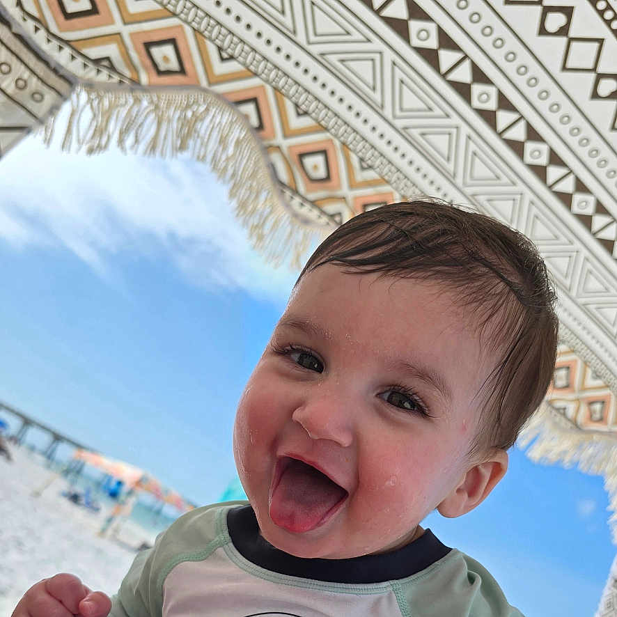 Arian is registered to the contest to win money with this photo: beach, blue_sky, casual_clothing, child, close_up, face, fun, happy, outdoor, patterned_umbrella, person, playful, smiling, summer, sunny, toddler, tongue_out, umbrella, vacation, wet_hair