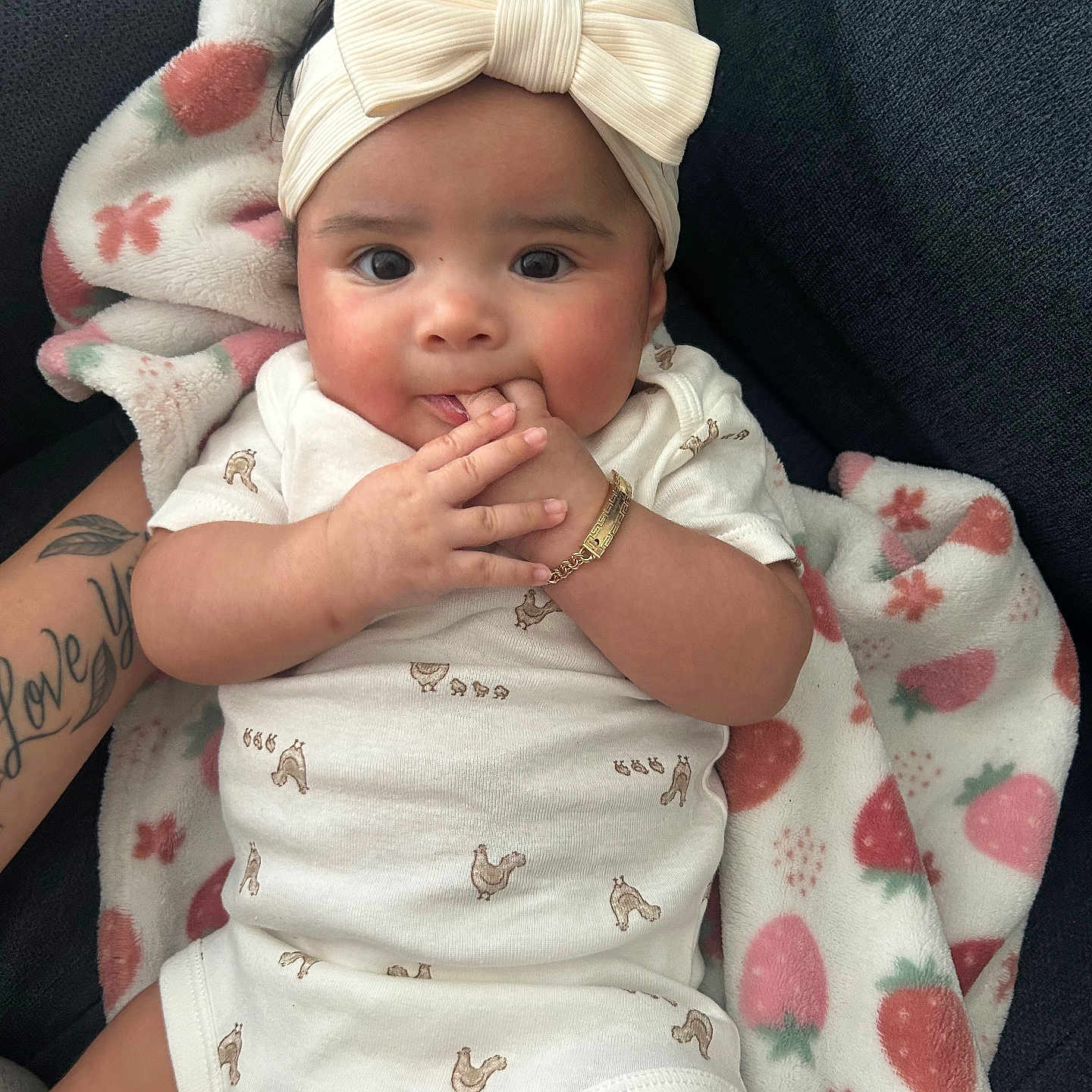 Leylani Ramos joined the competition — help win amazing prizes! baby, blanket, bow, chewing, chicken_print, child, couch, cute, face, fingers, hand, headband, indoors, infant, onesie, person, relaxed, soft_toy, strawberry_pattern, tattoo