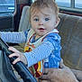 baby, child, steering_wheel, car_interior, hands, blue_eyes, bib, striped_clothing, seat, window, outdoor_view, vintage_car, person, supporting_hand, curious_expression, fingers, foot, house, trees, daylight