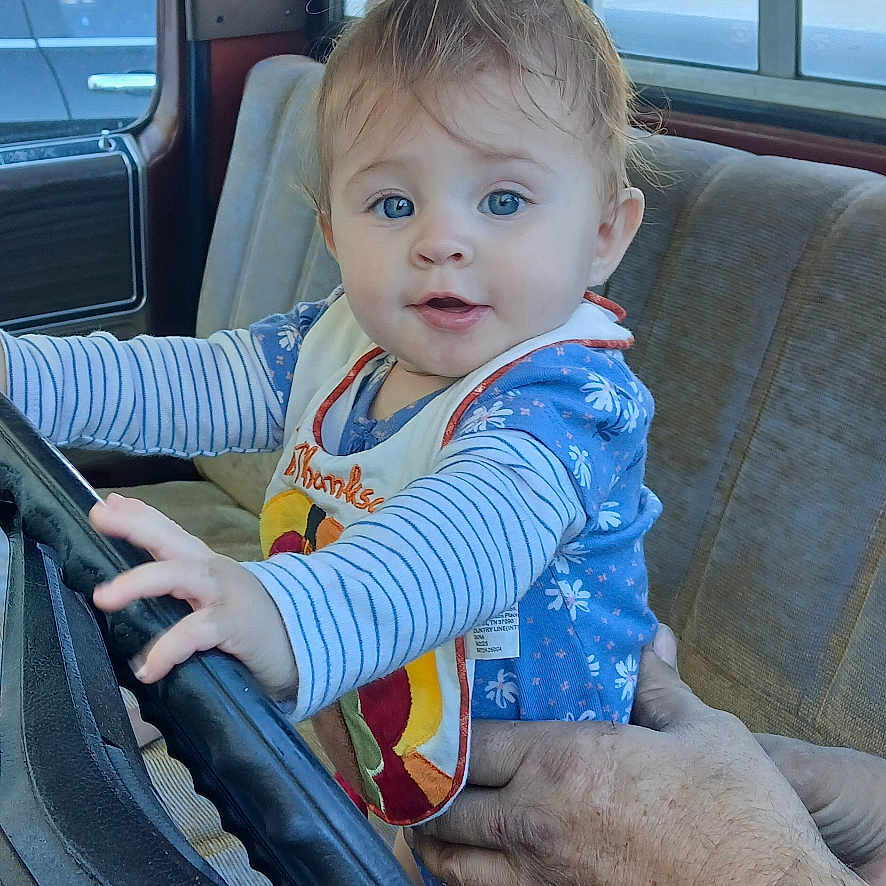 Ashlynn is registered to the contest to win money with this photo: baby, bib, blue_eyes, car_interior, child, curious_expression, daylight, fingers, foot, hands, house, outdoor_view, person, seat, steering_wheel, striped_clothing, supporting_hand, trees, vintage_car, window