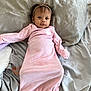baby, infant, pink_onesie, headband, pacifier, bed, gray_bedspread, lying_down, looking_at_camera, curious, soft_lighting, indoors, child, small_hands, cute, sleepwear, blanket, comfortable, portrait, young_child