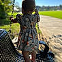 toddler, child, swing, bench, dress, floral_pattern, barefoot, outdoor, park, sunlight, grass, sidewalk, holding, chain, back_view, metal, summer, nature, shadow, play