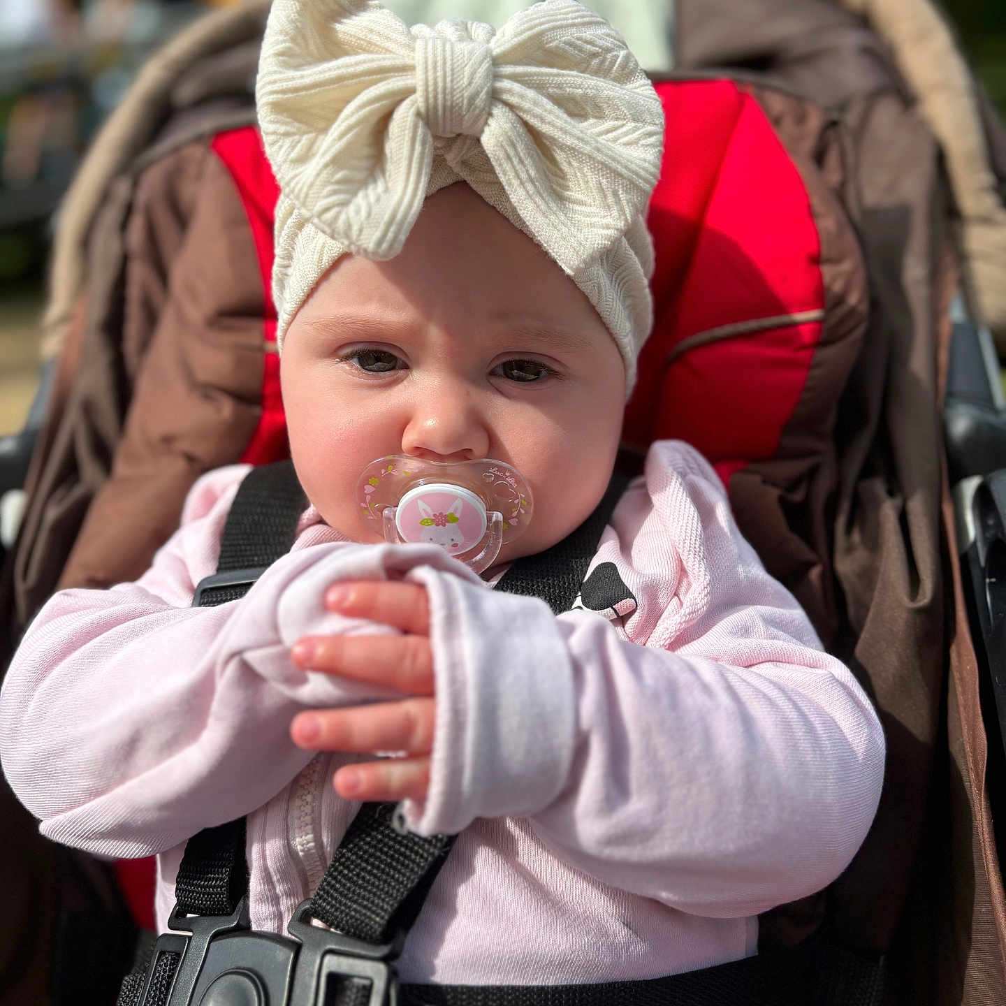 Inaya a rejoint le concours — aidez-le/la à gagner de superbes lots ! baby, black_straps, bow, brown_cushion, child, closeup, cute, daylight, face, hands, headband, infant, outdoor, pacifier, person, pink_clothing, portrait, red_cushion, seated, stroller