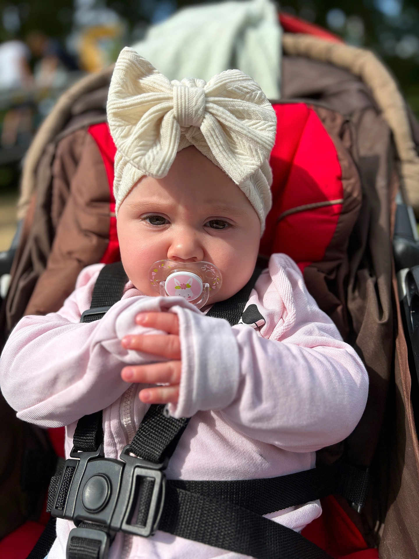 Inaya a rejoint le concours — aidez-le/la à gagner de superbes lots ! baby, pacifier, headband, bow, stroller, child, infant, pink_clothing, black_straps, red_cushion, brown_cushion, outdoor, daylight, portrait, closeup, cute, face, hands, seated, person