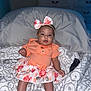 Addelyn joined the competition — help win amazing prizes! baby, infant, child, girl, headband, bow, orange_clothing, floral_skirt, bed, pillow, blanket, indoor, smiling, cute, person, young_child, patterned_bedding, happy, sitting, portrait
