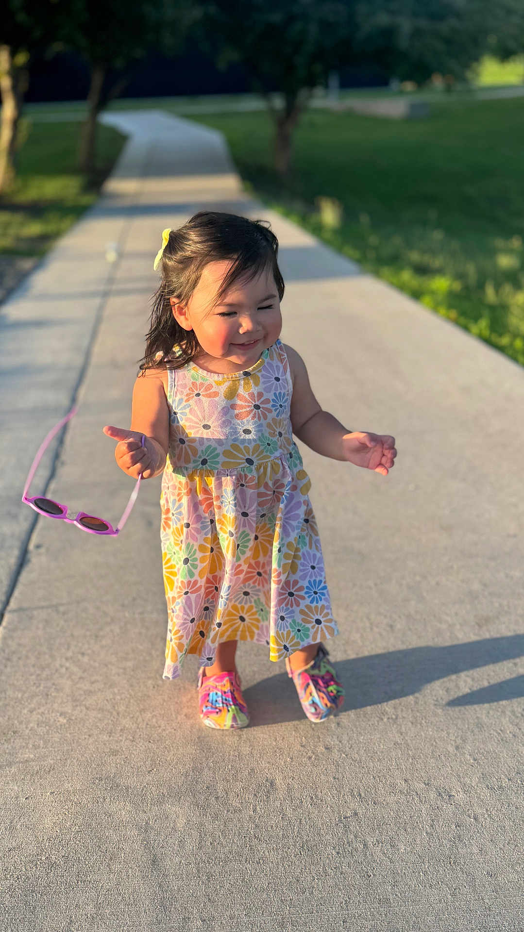 Milli is registered to the contest to win money with this photo: casual, child, colorful_dress, floral_pattern, girl, grass, happy, outdoor, person, playful, portrait, shoes, sidewalk, smiling, summer, sunglasses, sunlight, toddler, trees, walking