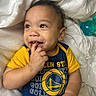 Royal is registered to the contest to win money with this photo: baby, child, infant, smiling, face, hand, blanket, clothing, blue, yellow, golden_state_warriors, sportswear, cute, happy, lying_down, indoor, soft, playful, fingers, toddler
