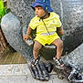 bucket_hat, child, concrete, curious, daylight, expression, footwear, gorilla, hand, hat, outdoor, playful, sandals, sculpture, side_view, sitting, statue, toddler, urban, yellow_clothing