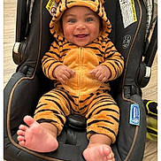 Antonio is registered to the contest to win money with this photo: baby, child, car_seat, tiger_onesie, smiling, cute, infant, hooded_clothing, footwear_bare, indoor, happy, person, plush, cozy, seated, wood_floor, orange, black_stripes, adorable, small_feet