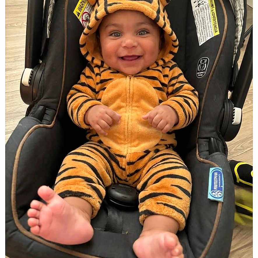 Antonio is registered to the contest to win money with this photo: baby, child, car_seat, tiger_onesie, smiling, cute, infant, hooded_clothing, footwear_bare, indoor, happy, person, plush, cozy, seated, wood_floor, orange, black_stripes, adorable, small_feet