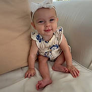 Madison is registered to the contest to win money with this photo: baby, child, infant, smiling, headband, bow, floral_clothing, sitting, couch, pillow, indoor, window, natural_light, skin, face, hands, feet, white, beige, cozy