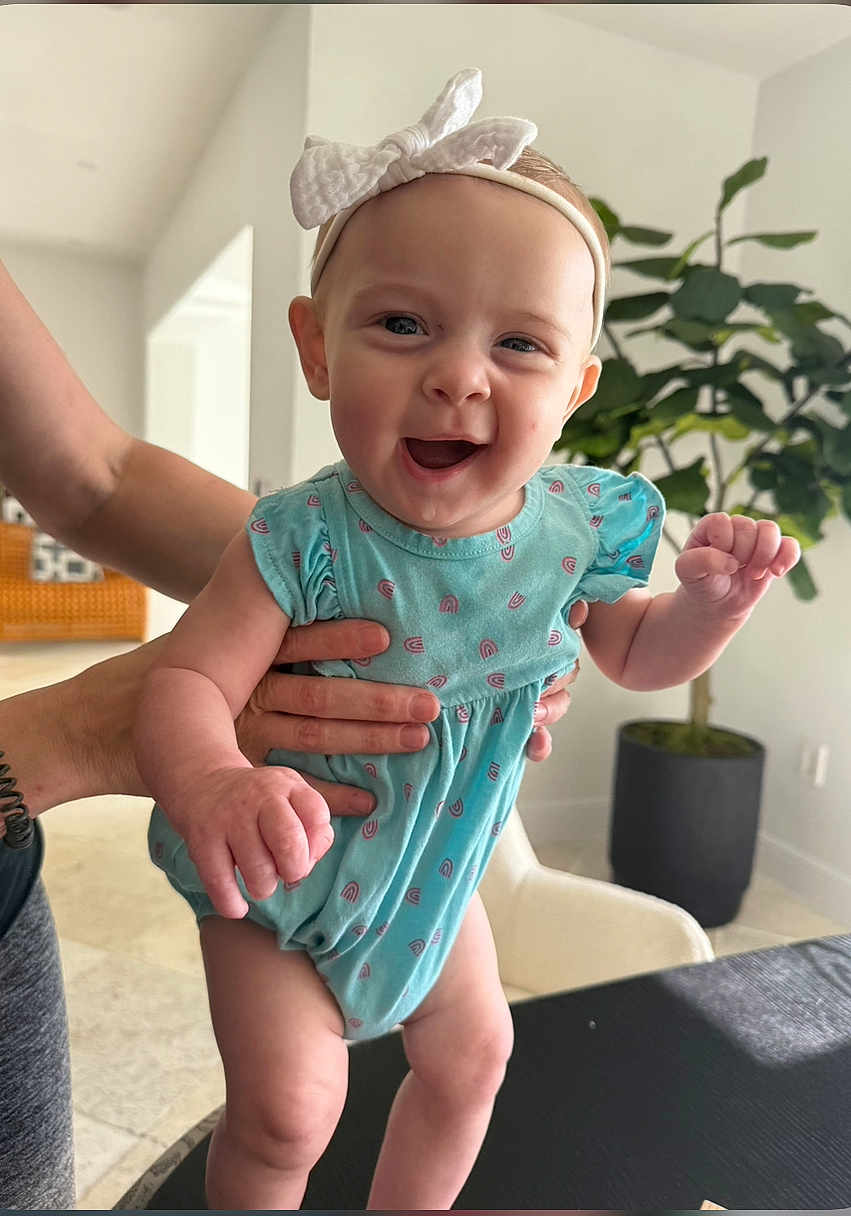 Madison joined the competition — help win amazing prizes! baby, child, smiling, happy, onesie, headband, bow, indoor, person, hands, plant, potted_plant, furniture, table, arm, face, cute, expression, infant, light