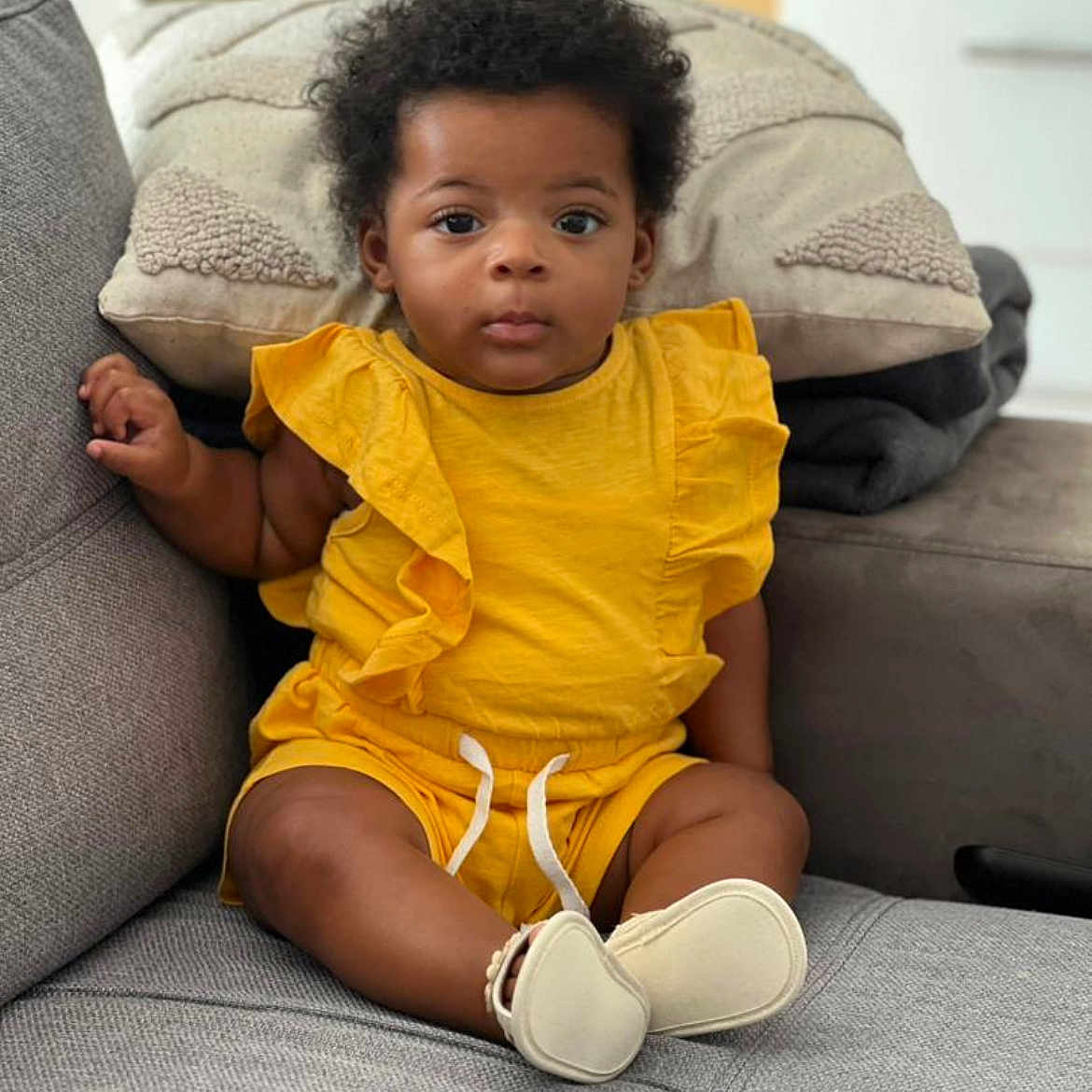 Eden participe au concours pour gagner de l'argent avec cette photo : baby, casual, child, comfort, couch, cozy, curly_hair, cute, expression, footwear, furniture, home, indoor, infant, person, pillow, portrait, relaxing, sitting, yellow_clothing