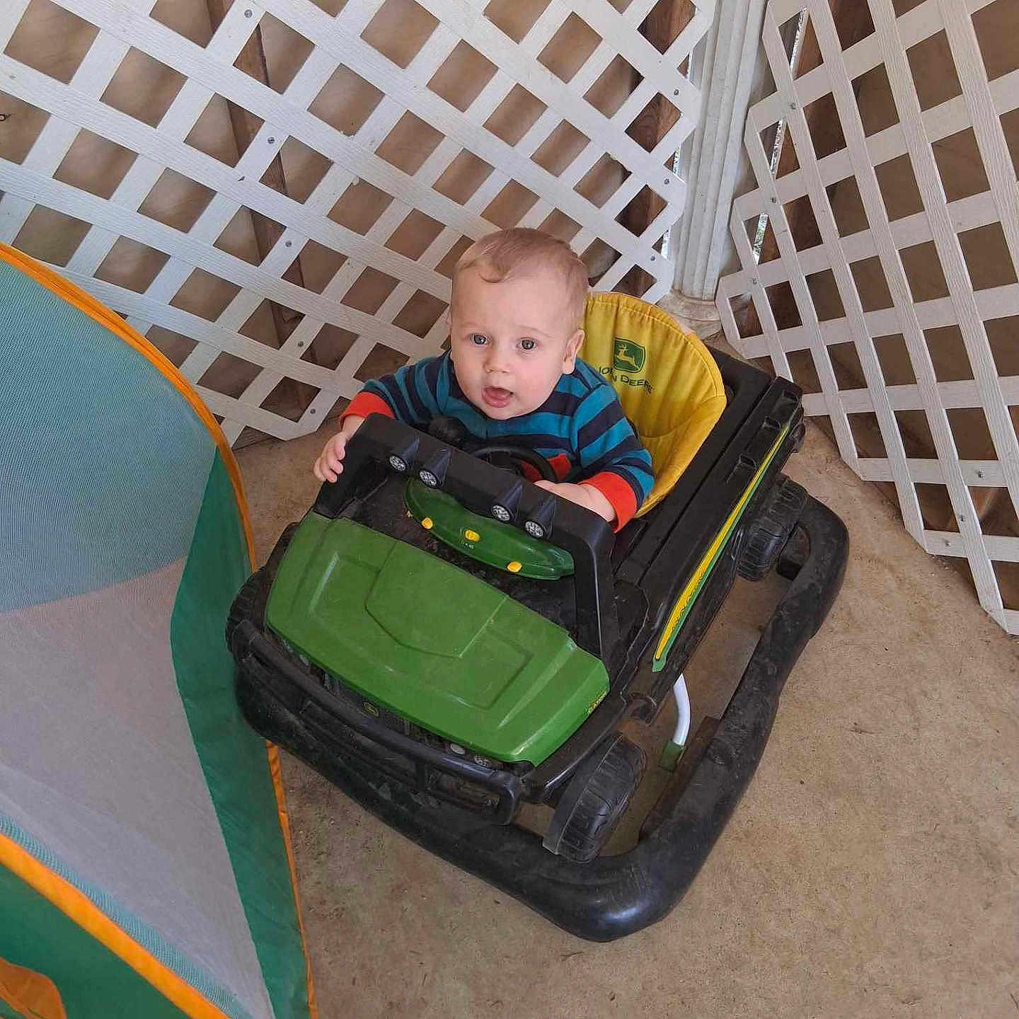 Zach is registered to the contest to win money with this photo: baby, child, curious, cute, face, floor, head, indoor, infant, john_deere, lattice_fence, person, plastic, play_tent, playtime, seated, small, striped_shirt, toy_car, wheels