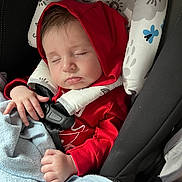 Icarus is registered to the contest to win money with this photo: baby, blanket, car_seat, child, clothing, cozy, cute, face, hand, headrest, indoors, infant, pattern, peaceful, red_hoodie, resting, safety_harness, sleeping, toddler, young_child
