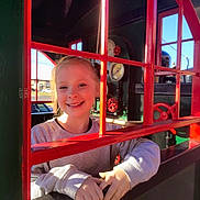 Harper is registered to the contest to win money with this photo: casual_clothing, child, daylight, face, girl, hands, happy, metal, outdoor, play, portrait, red_bars, smile, sunlight, sweater, train_cabin, vehicle, vintage, window, young_person