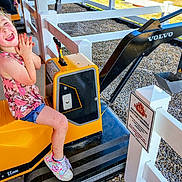 Hannah is registered to the contest to win money with this photo: casual, child, daylight, excavator, fence, fun, girl, gravel, happy, joy, outdoor, playground, ride, shoes, shorts, smiling, summer, tank_top, toy, yellow