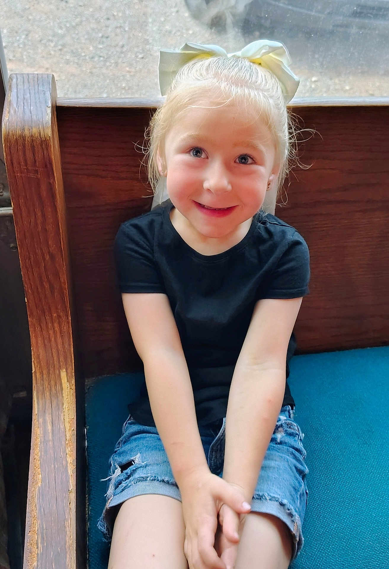 Hannah joined the competition — help win amazing prizes! black_shirt, blue_cushion, bow, casual_clothing, child, cute, denim_shorts, face, girl, hair, hands, happy, indoors, light, person, portrait, sitting, smile, wooden_bench, young
