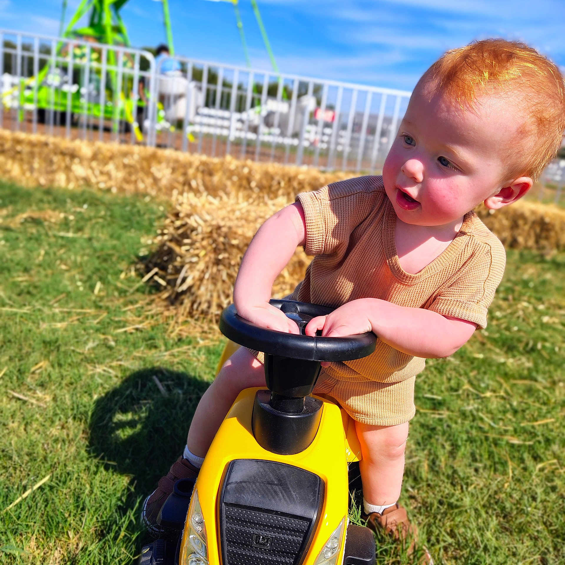 Henry joined the competition — help win amazing prizes! toddler, child, toy_car, yellow, grass, outdoor, sunny, red_hair, rosy_cheeks, playground, hay_bales, steering_wheel, shorts, shirt, shoes, summer, daytime, curious, playing, fun