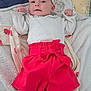 baby, infant, child, clothing, headband, blanket, fabric, footwear, red_shorts, white_top, bed, soft_texture, cute, portrait, lying_down, indoors, comfort, pose, person, face