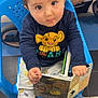 blond_hair, blue_chair, book, boots, child, clothing, curious, cute, floor_tiles, holding, indoor, pants, person, playful, reading, seated, simba_shirt, stroller, toddler, toy