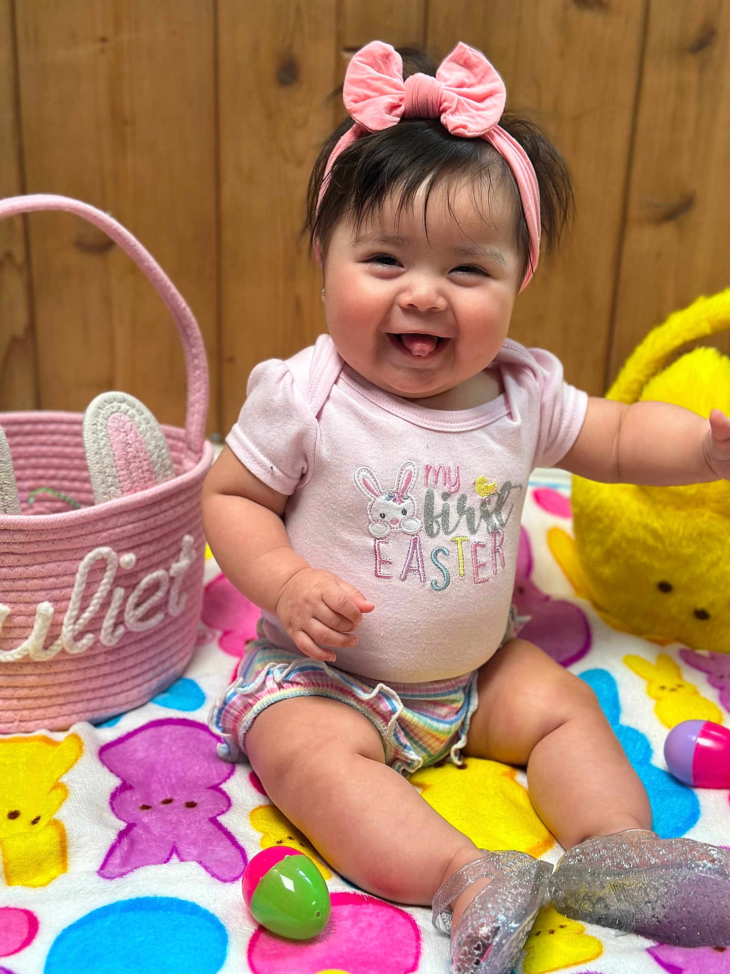 Juliet is registered to the contest to win money with this photo: baby, girl, pink_bow, headband, shirt, easter, colorful_blanket, bunny_pattern, easter_eggs, basket, smiling, tongue_out, wooden_background, sitting, cute, shorts, sparkly_shoes, indoor, happy, celebration