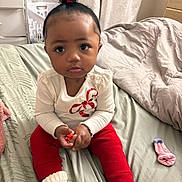 Journeerose is registered to the contest to win money with this photo: toddler, child, indoor, bed, blanket, clothing, socks, red_pants, white_shirt, curly_hair, hair_tie, face, person, cute, serious_expression, room, fabric, knit_socks, pink_sock, handheld_object