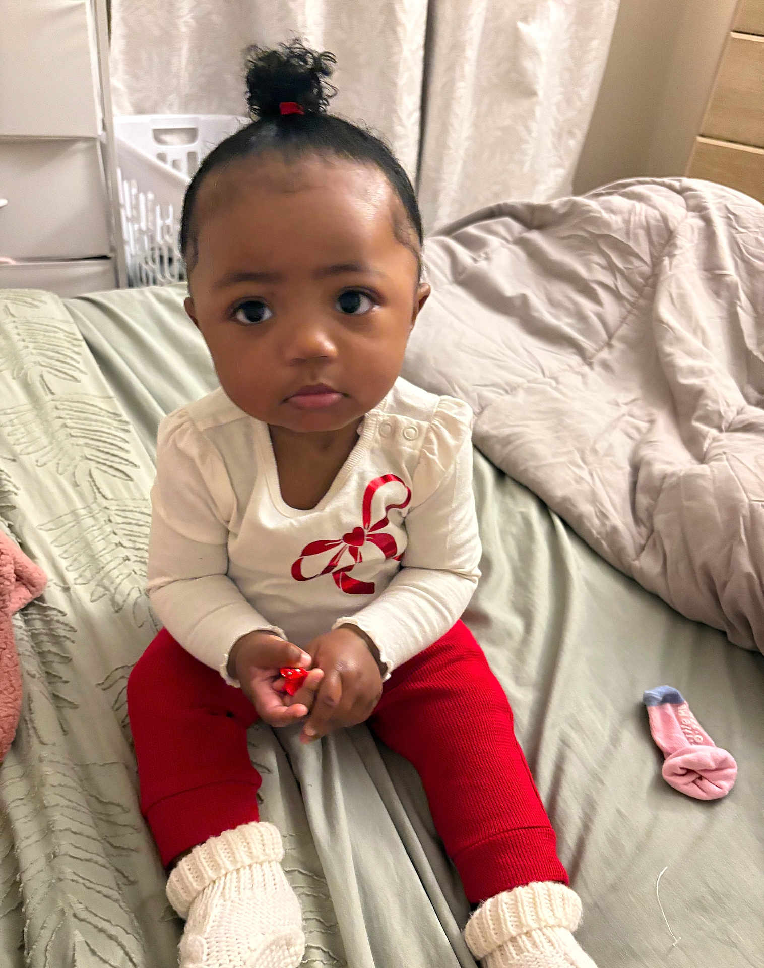 Journeerose is registered to the contest to win money with this photo: toddler, child, indoor, bed, blanket, clothing, socks, red_pants, white_shirt, curly_hair, hair_tie, face, person, cute, serious_expression, room, fabric, knit_socks, pink_sock, handheld_object