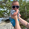 Xander is registered to the contest to win money with this photo: baby, child, sunglasses, beach, trees, water, person, hand, sky, nature, summer, outdoor, holding, clothing, tattoo, playful, cute, sunshine, casual, portrait