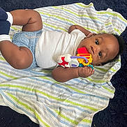 Makari is registered to the contest to win money with this photo: baby, infant, toy, blanket, striped_blanket, lying_down, curious, chewing, clothing, white_shirt, blue_diaper, socks, indoor, carpet, black_carpet, hand, face, eyes, cute, child