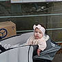 baby, child, stroller, headband, pink_bow, knitted_sweater, outdoor, cardboard_box, bus_stop, signage, gray_stroller, person, infant, cute, seated, urban, sidewalk, casual_clothing, young_child, portrait