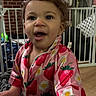baby_gate, blanket, brick_wall, child, clothing, curly_hair, door, flower_pattern, furniture, hand, happy, home, indoor, person, pink_shirt, portrait, smiling, strawberry_pattern, toddler, wooden_floor