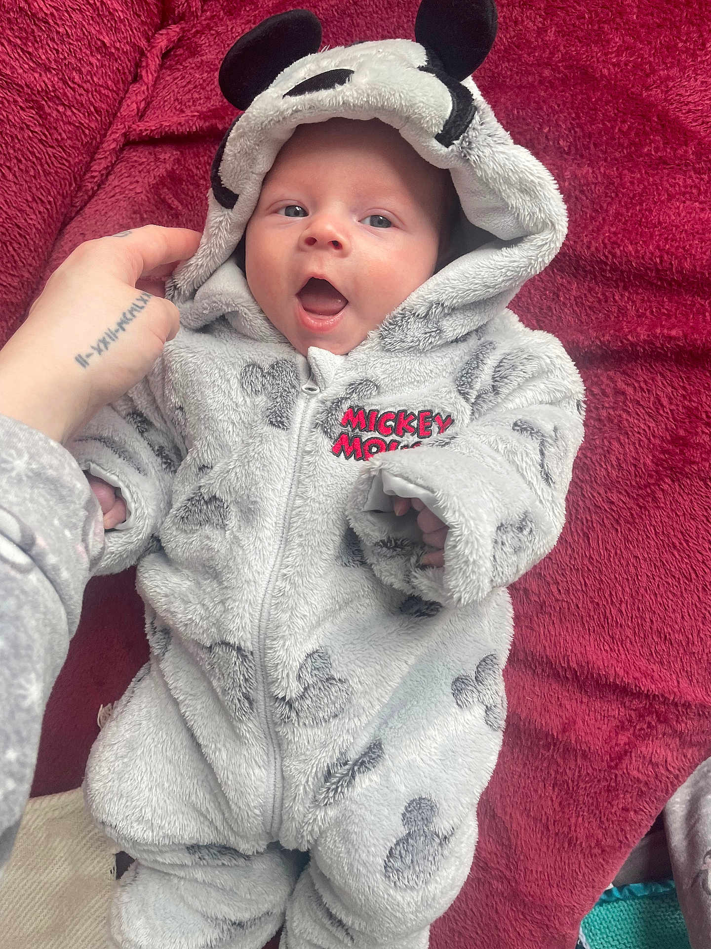 Roman is registered to the contest to win money with this photo: baby, infant, mickey_mouse, onesie, hood, ears, hand, red_blanket, smile, happy, clothing, soft, cute, lying_down, portrait, child, baby_clothes, indoor, person, zipped_jumpsuit