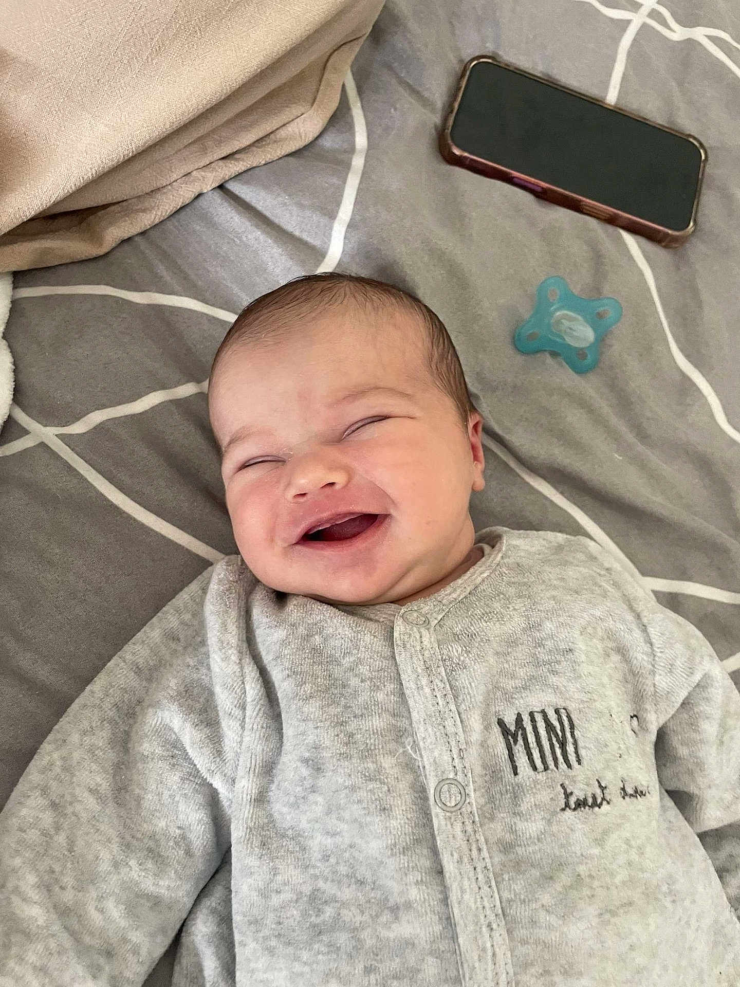 Emmy a rejoint le concours — aidez-le/la à gagner de superbes lots ! baby, infant, smiling, laughing, onesie, grey_clothing, bed, pacifier, phone, happy, face, person, lying_down, indoor, cute, newborn, child, fabric, text_on_clothing, closeup