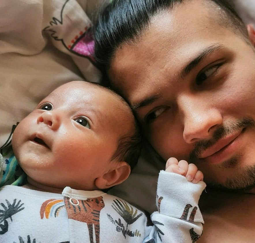 Teddy joined the competition — help win amazing prizes! baby, infant, man, adult, face, portrait, closeup, bed, blanket, onesie, animal_print, smile, mustache, goatee, skin, head, hand, lying_down, indoors, family