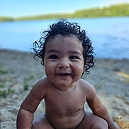 Romeo is registered to the contest to win money with this photo: baby, child, curly_hair, smiling, sitting, lake, water, sand, outdoor, nature, happy, portrait, young_child, bare_chest, sunlight, summer, playful, innocence, cute, closeup