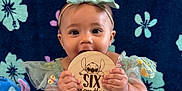 Marilyn is registered to the contest to win money with this photo: baby, child, infant, green_dress, headband, plush_toy, stitch_character, floral_pattern, blue_blanket, wooden_plaque, six_months, cute, smiling, sitting, indoors, portrait, playful, decorative, soft_toys, happy